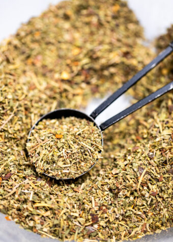 A measuring spoon scooping Italian Seasoning from a bowl of herbs and spices.