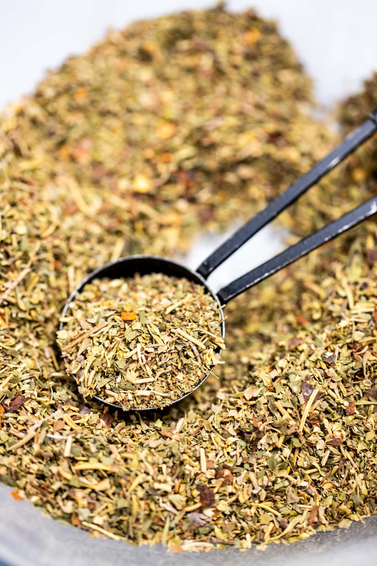 A measuring spoon scooping Italian Seasoning from a bowl of herbs and spices.