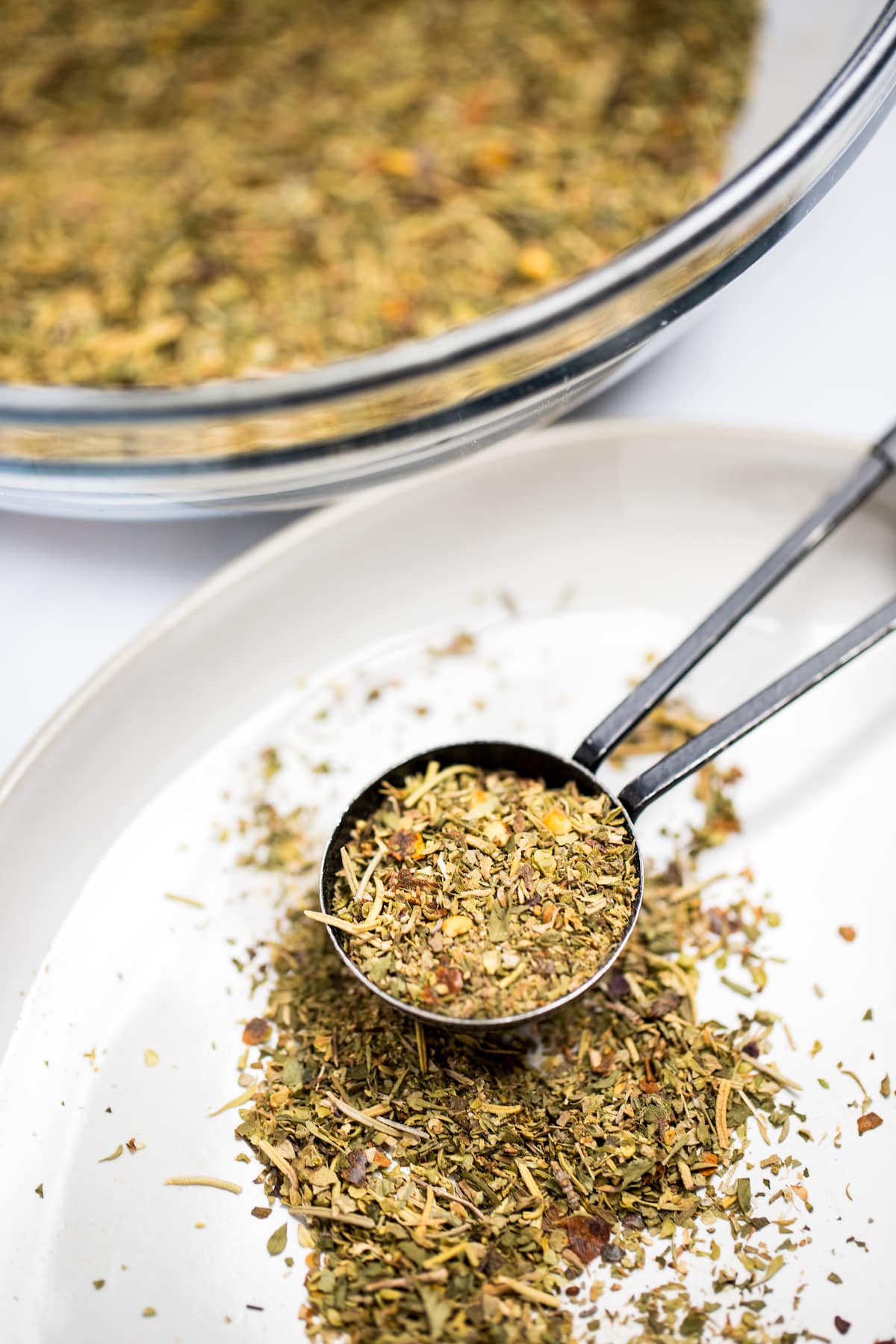 A measuring spoon with a scoop of Italian Seasoning from a bowl of herbs and spices, on a plate with the spice blend spilling out.