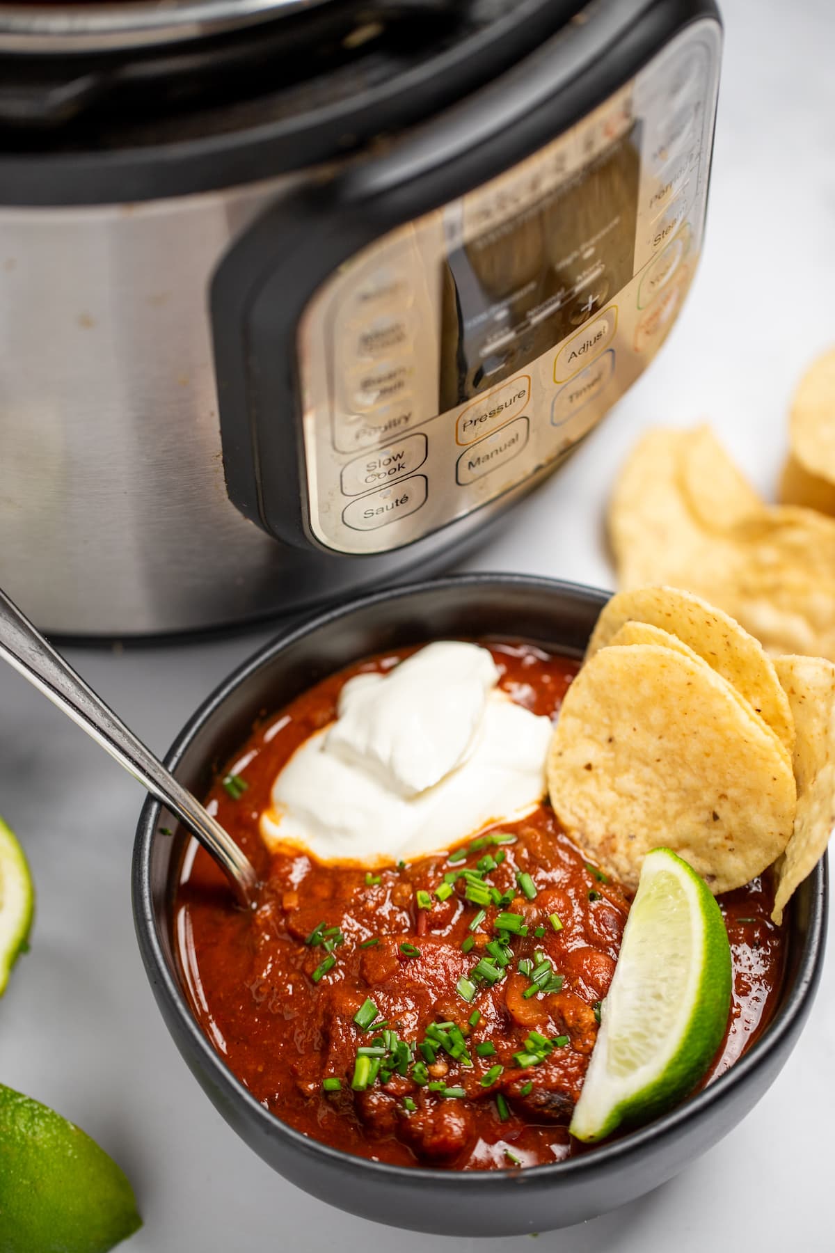 A bowl of chili with a spoon in it, topped with chopped chives, a lime wedge, and round tortilla chips, next to more lime wedges and tortilla chips, in front of an instant pot.