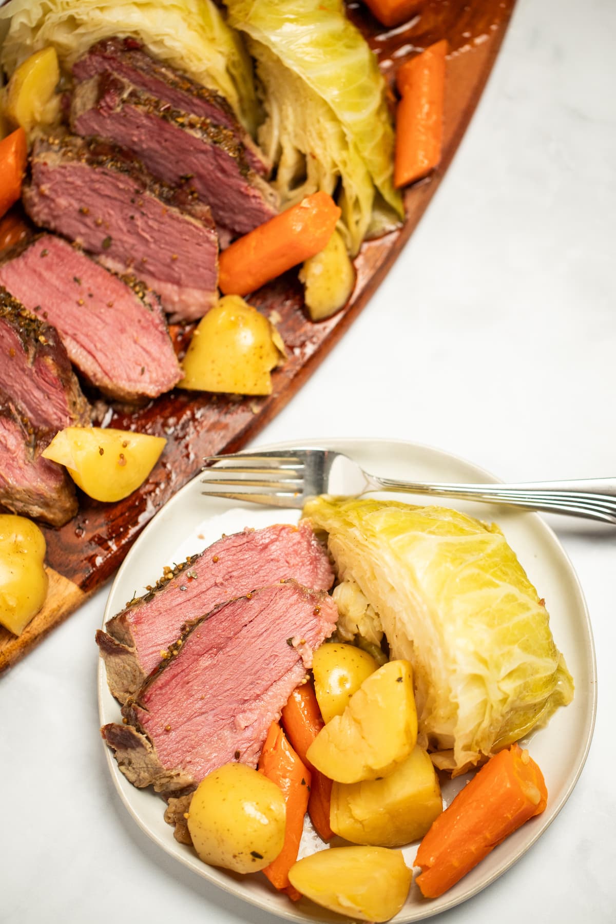 Instant pot corned beef with potatoes and cabbage on a wooden cutting board, next to a plate with corned beef, potatoes, cabbage, and carrots on it, with a fork on a table.