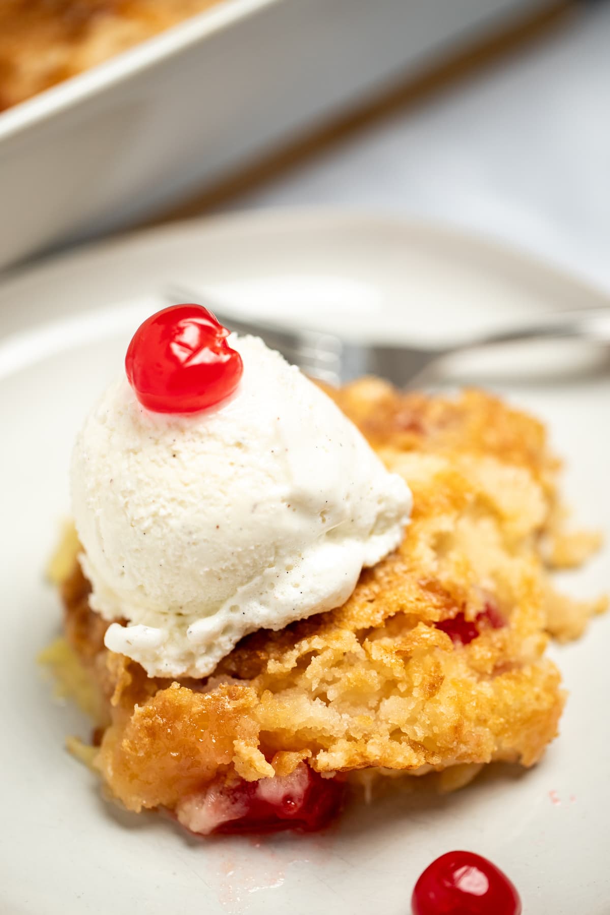 A piece of pineapple dump cake on a plate topped with ice cream and maraschino cherries.