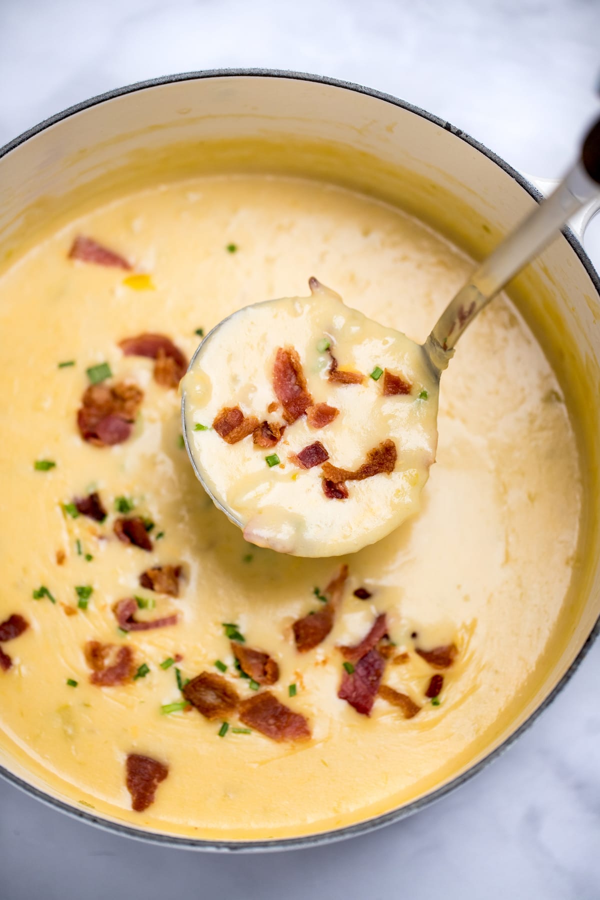 A dutch oven full of potato cheese soup topped with bacon and chives, with a ladle scooping up soup.