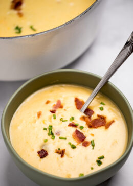 A bowl of potato cheese soup topped with chives and bacon with a spoon in the bowl, in front of a dutch oven full of soup.