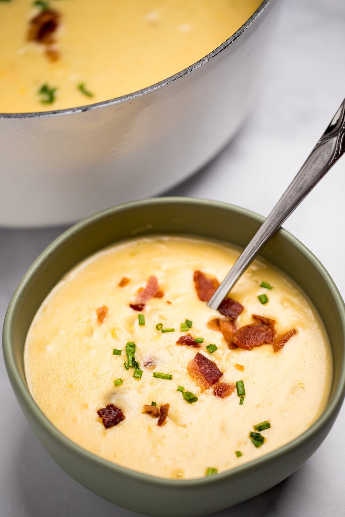 A bowl of potato cheese soup topped with chives and bacon with a spoon in the bowl, in front of a dutch oven full of soup.