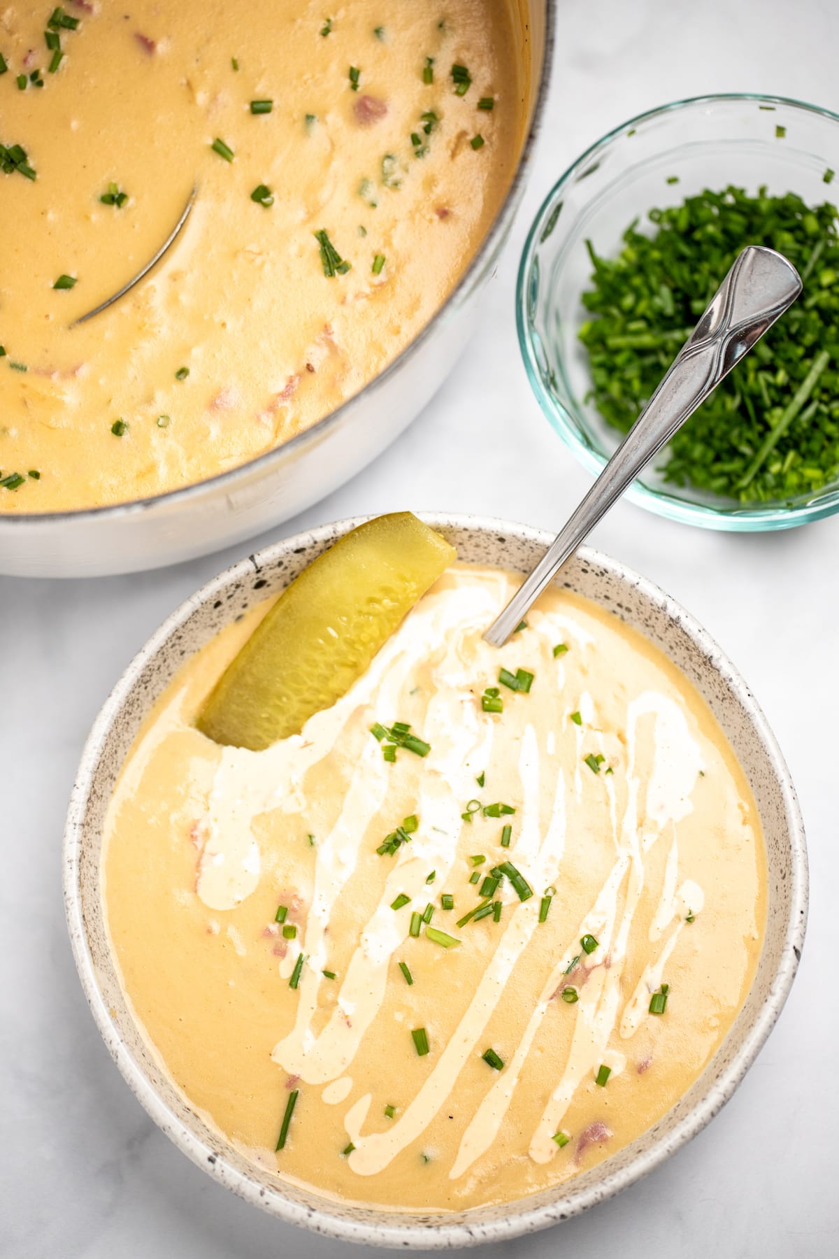 A spoon in a bowl of reuben soup on a table, topped with a pickle and a drizzle of Thousand Island dressing and chopped chives, in front of a dutch oven of soup and a small bowl of chives.