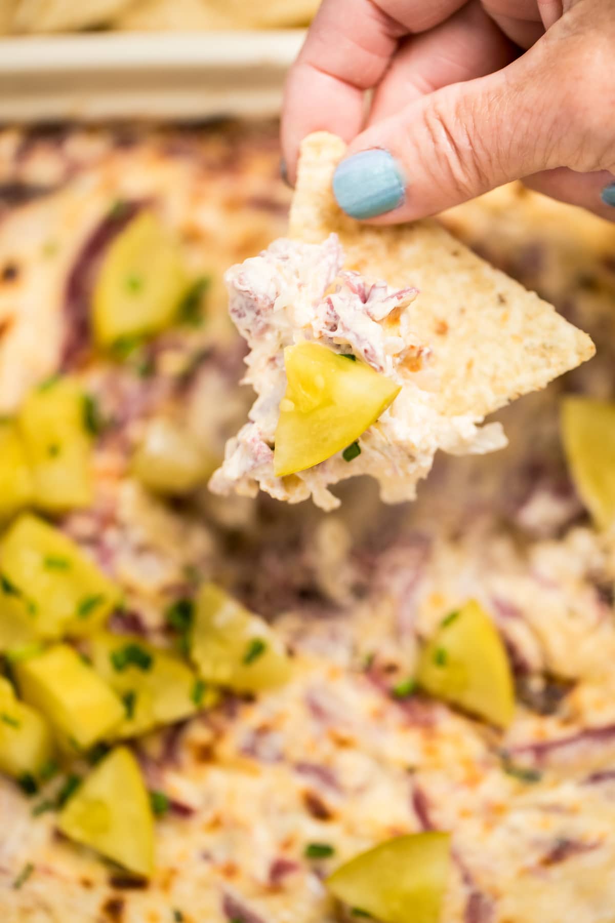 A hand holding a tortilla chip with reuben dip and a pickle on it, over a dish of reuben dip topped with pickles.
