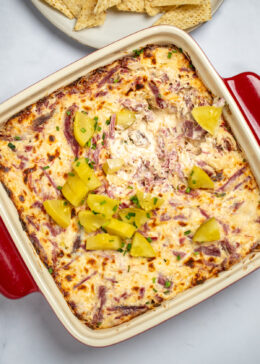 Reuben dip topped with pickles in a baking dish on a table, with a plate of chips behind it.