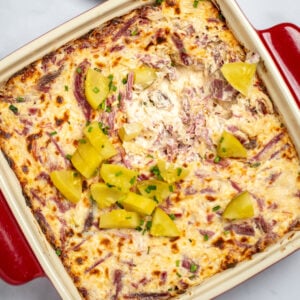 Reuben dip topped with pickles in a baking dish on a table, with a plate of chips behind it.