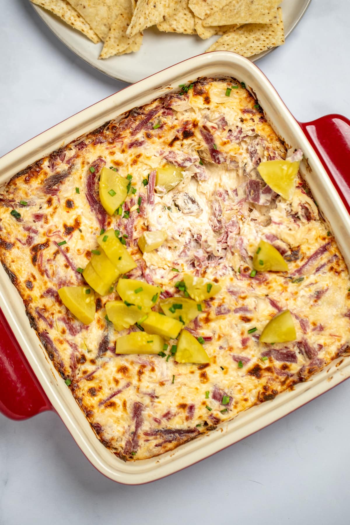 Reuben dip topped with pickles in a baking dish on a table, with a plate of chips behind it.