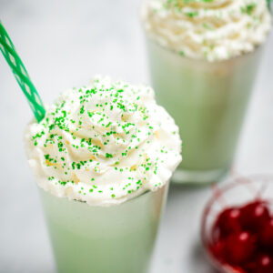 A tall glass with shamrock shake topped with whipped cream and sprinkles, next to a bowl of maraschino cherries. There is another shake in the background, and they both have straws.