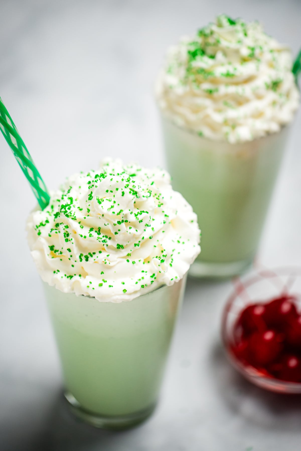 A tall glass with shamrock shake topped with whipped cream and sprinkles, next to a bowl of maraschino cherries. There is another shake in the background, and they both have straws.