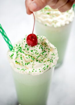 A tall glass with shamrock shake topped with whipped cream, sprinkles, and a hand lowering a cherry on top. There is another shake in the background, and they both have straws.