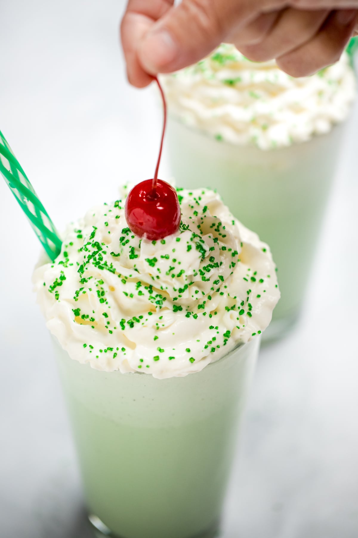 A tall glass with shamrock shake topped with whipped cream, sprinkles, and a hand lowering a cherry on top. There is another shake in the background, and they both have straws.