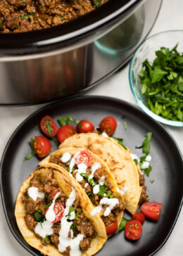 Three beef tacos on a plate with cherry tomatoes, cilantro, and sour cream drizzled on top, in front of a slow cooker with slow cooker taco meat.