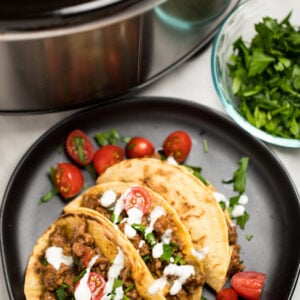 Three beef tacos on a plate with cherry tomatoes, cilantro, and sour cream drizzled on top, in front of a slow cooker with slow cooker taco meat.