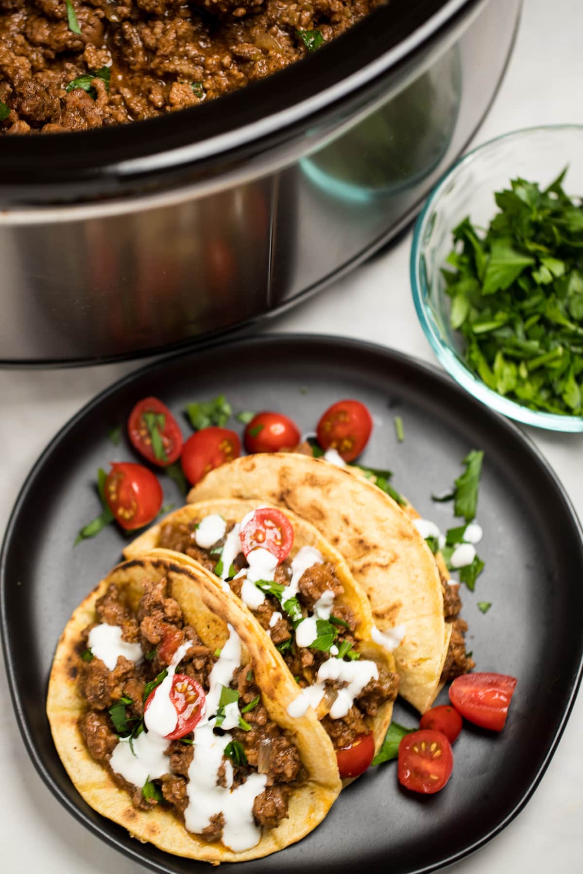 Three beef tacos on a plate with cherry tomatoes, cilantro, and sour cream drizzled on top, in front of a slow cooker with slow cooker taco meat.