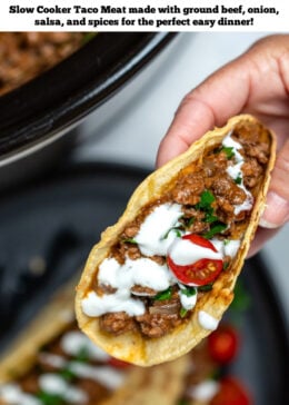 Pinterest pin with a hand holding a beef taco drizzled with sour cream and cherry tomatoes above a plate of tacos, in front of a slow cooker full of slow cooker taco meat.