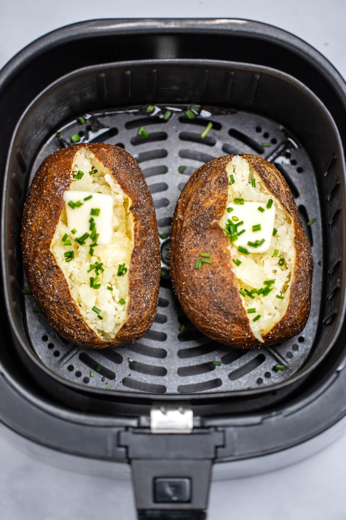 Two fully cooked baked potatoes topped with butter, chives, and sour cream sitting in an air fryer basket.