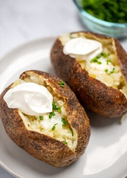 Two baked potatoes on a plate topped with butter, chives, and sour cream.