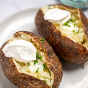 Two baked potatoes on a plate topped with butter, chives, and sour cream.