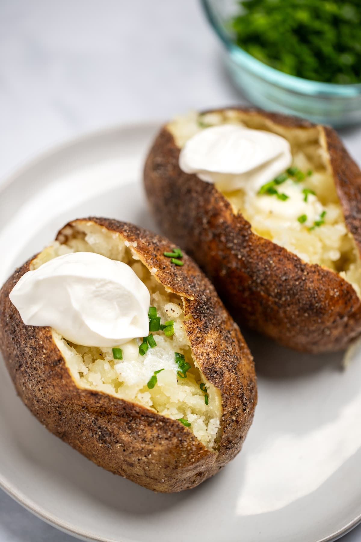 Two baked potatoes on a plate topped with butter, chives, and sour cream.