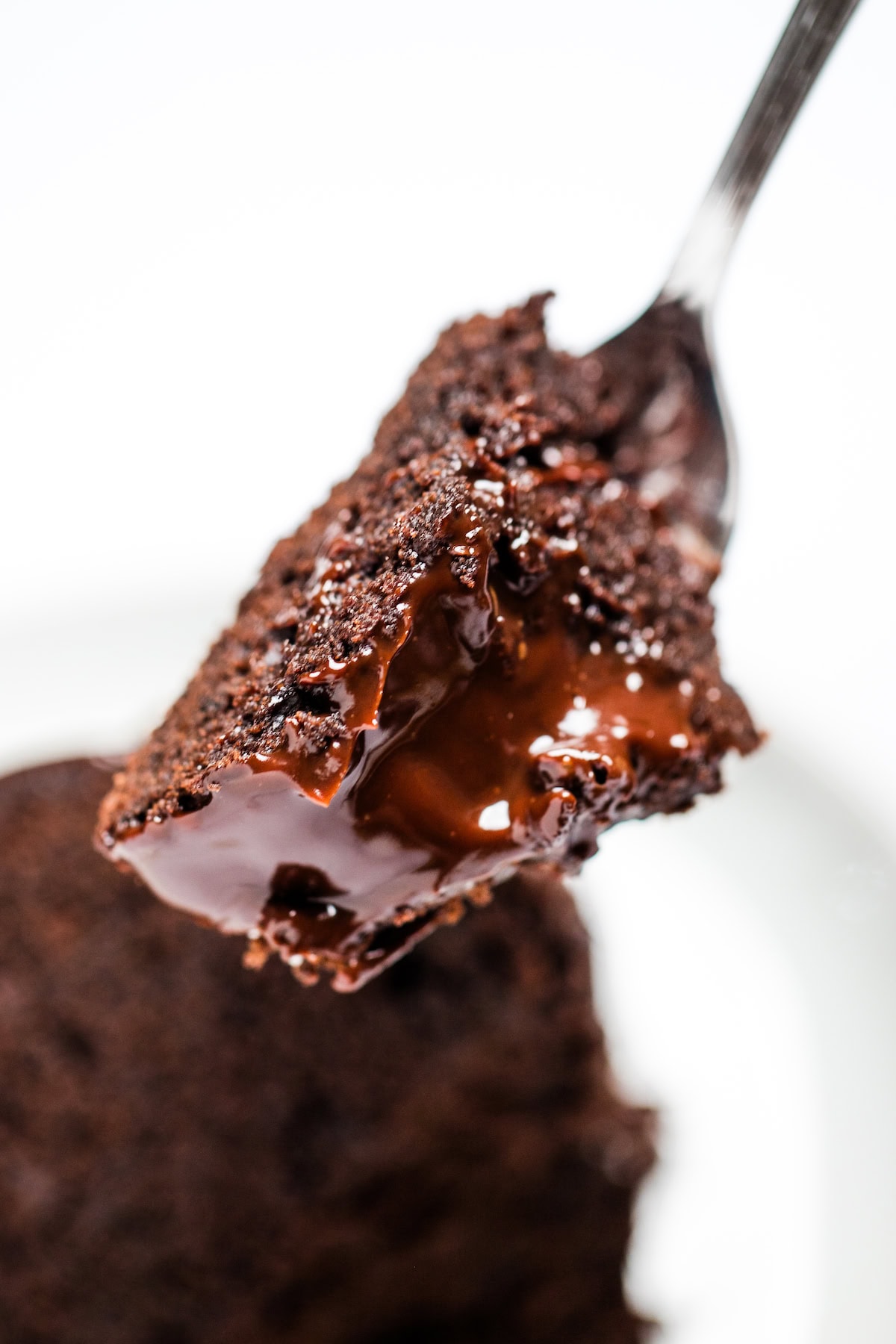A closeup of a fork full of chocolate brownie cake with a creamy chocolate ganache on top, with a piece of cake in the background.