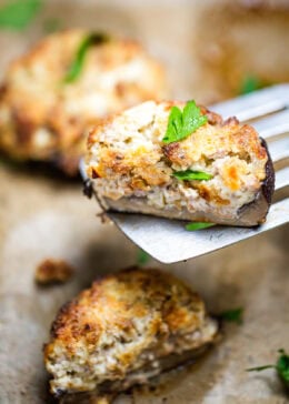 A sheet pan of stuffed portobello mushrooms with a spatula lifting up half a mushroom topped with fresh chopped parsley.