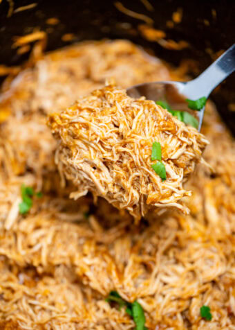 A slow cooker with saucy shredded BBQ chicken and a spoon lifting up the chicken.