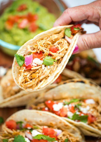 A hand holding a taco in a corn tortilla topped with tomato, red onion, queso fresco, and cilantro, above a cutting board with more tacos and a bowl of guacamole.