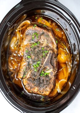 A fully cooked pork roast in a slow cooker with broth and onions, topped with fresh parsley.