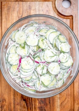 A glass mixing bowl on a cutting board full of creamy cucumber salad with red onions topped with fresh dill.