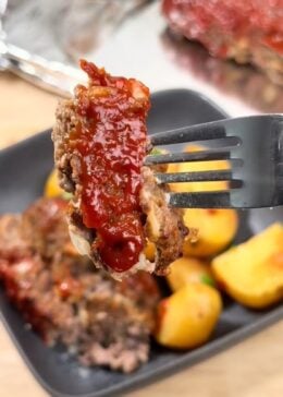 A fork holding a piece of meatloaf over a plate of crockpot meatloaf and potatoes, with a sheet pan wrapped in tinfoil and the whole meatloaf behind it.
