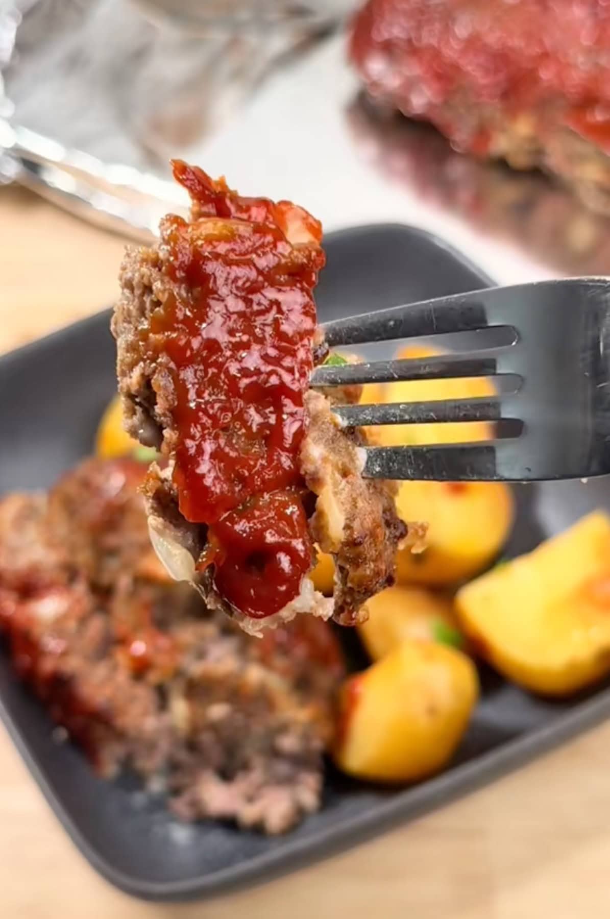 A fork holding a piece of meatloaf over a plate of crockpot meatloaf and potatoes, with a sheet pan wrapped in tinfoil and the whole meatloaf behind it.