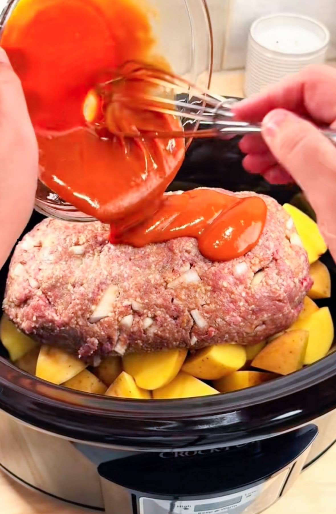 A slow cooker with quartered potatoes, with a raw meatloaf on top with glaze being poured from a bowl on top of the meatloaf.