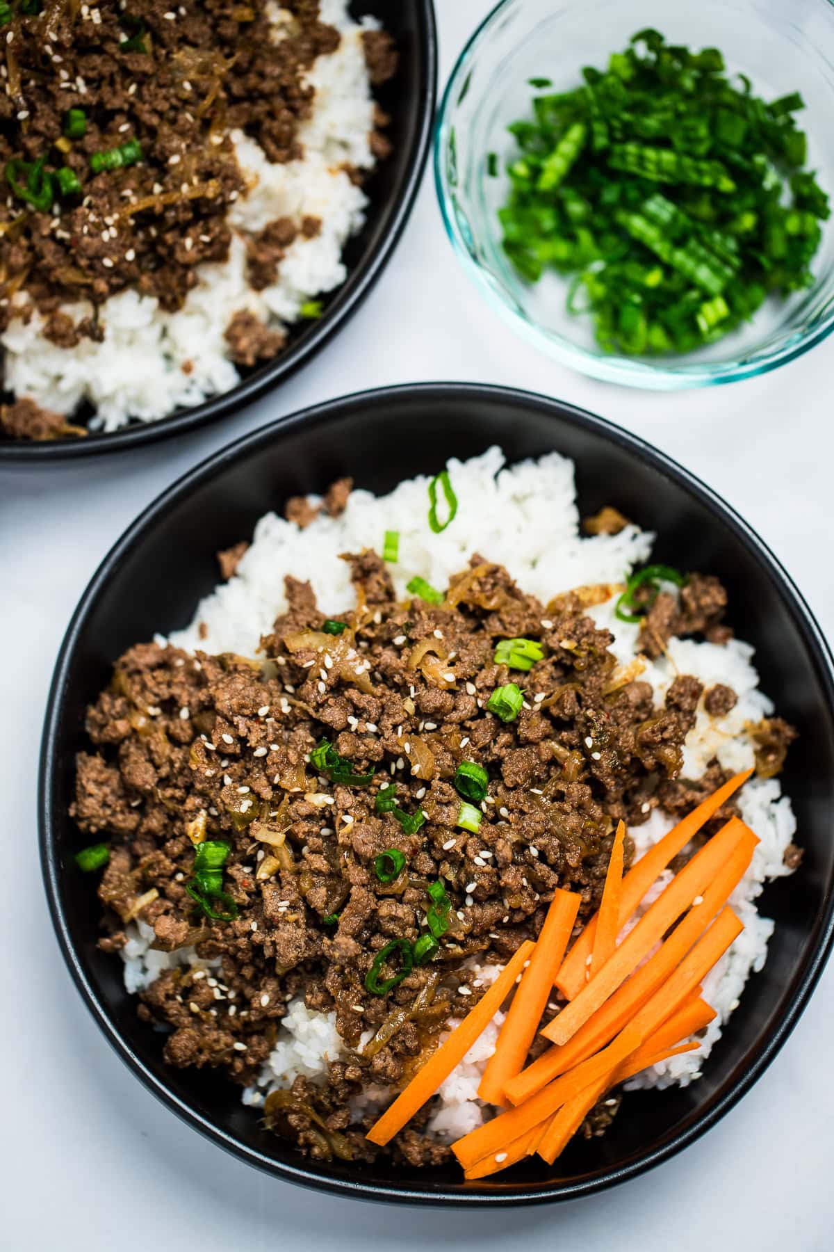 A pinterest pin with a bowl of rice topped with seasoned beef with raw carrot sticks and green onions, with another bowl and green onions in the background.