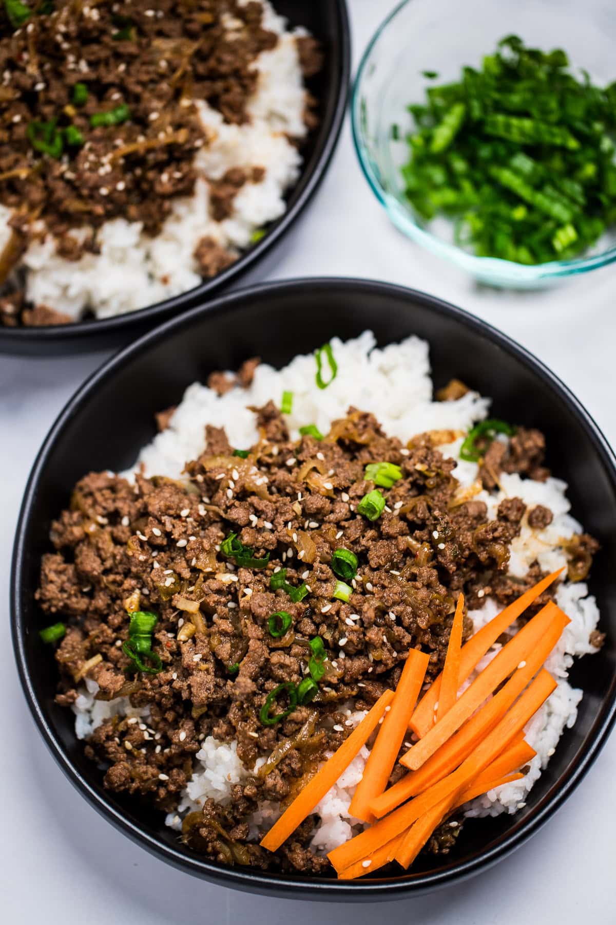 A pinterest pin with a bowl of rice topped with seasoned beef with raw carrot sticks and green onions, with another bowl and green onions in the background.