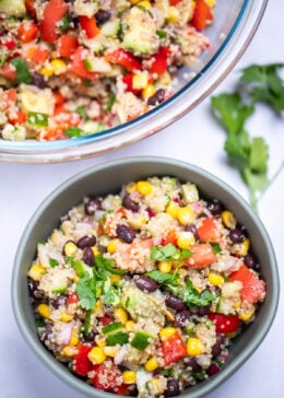 A bowl of black bean quinoa salad topped with fresh cilantro, with a larger glass bowl of salad in the background. A sprig of cilantro rests next to the smaller bowl on the table.