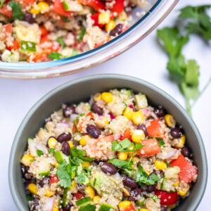 A bowl of black bean quinoa salad topped with fresh cilantro, with a larger glass bowl of salad in the background. A sprig of cilantro rests next to the smaller bowl on the table.