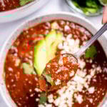 A spoon lifting black bean soup from a bowl topped with queso fresco, cilantro leaves, and sliced avocado, with a bowl of cilantro and lime wedges, and a dutch oven of soup in the background.