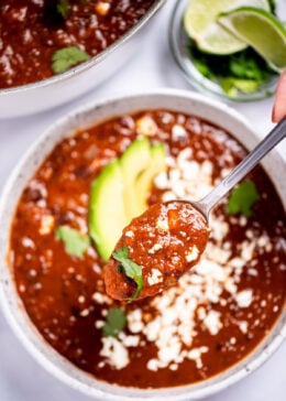 A spoon lifting black bean soup from a bowl topped with queso fresco, cilantro leaves, and sliced avocado, with a bowl of cilantro and lime wedges, and a dutch oven of soup in the background.