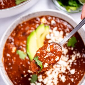 A spoon lifting black bean soup from a bowl topped with queso fresco, cilantro leaves, and sliced avocado, with a bowl of cilantro and lime wedges, and a dutch oven of soup in the background.