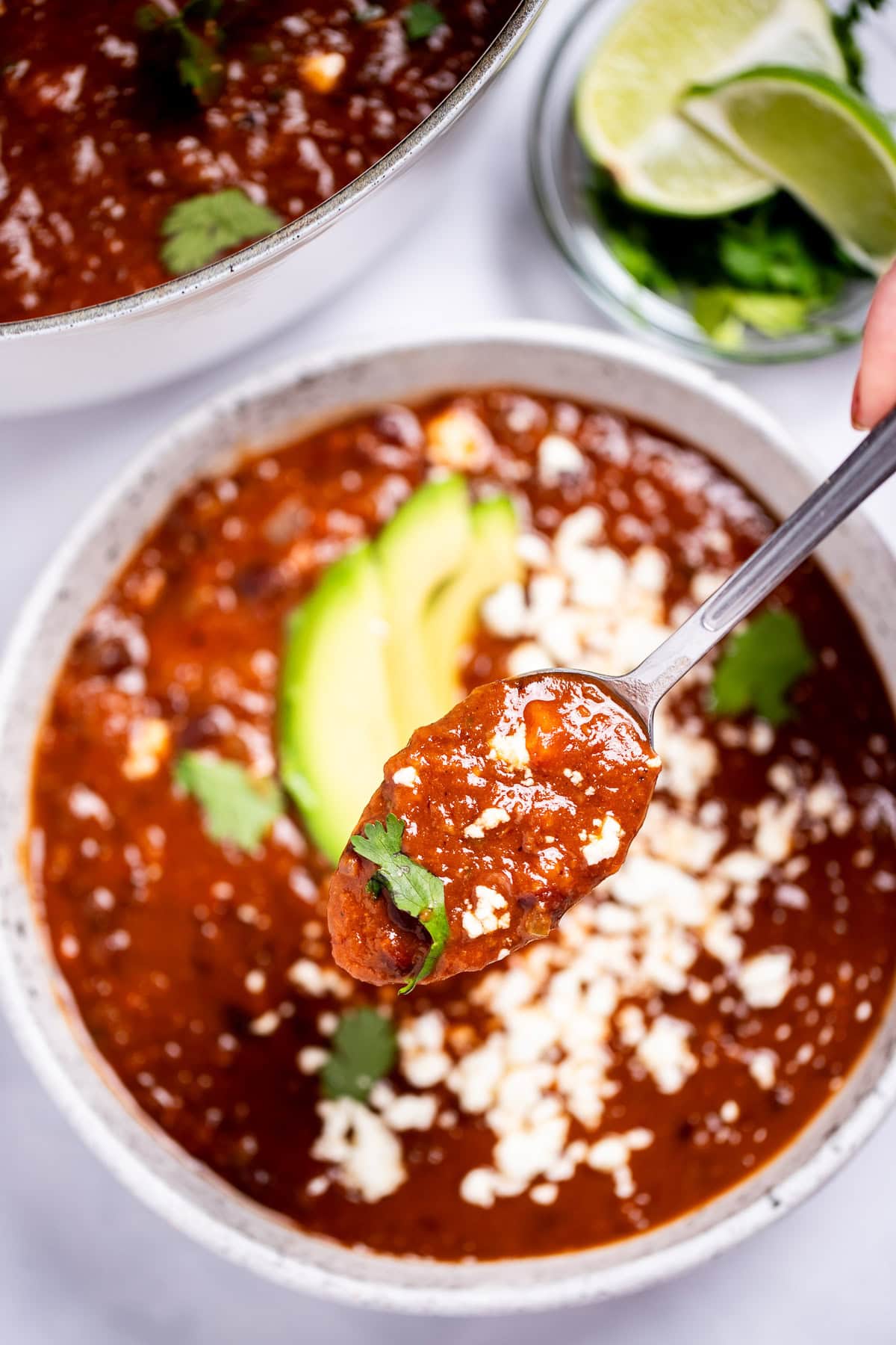A spoon lifting black bean soup from a bowl topped with queso fresco, cilantro leaves, and sliced avocado, with a bowl of cilantro and lime wedges, and a dutch oven of soup in the background.