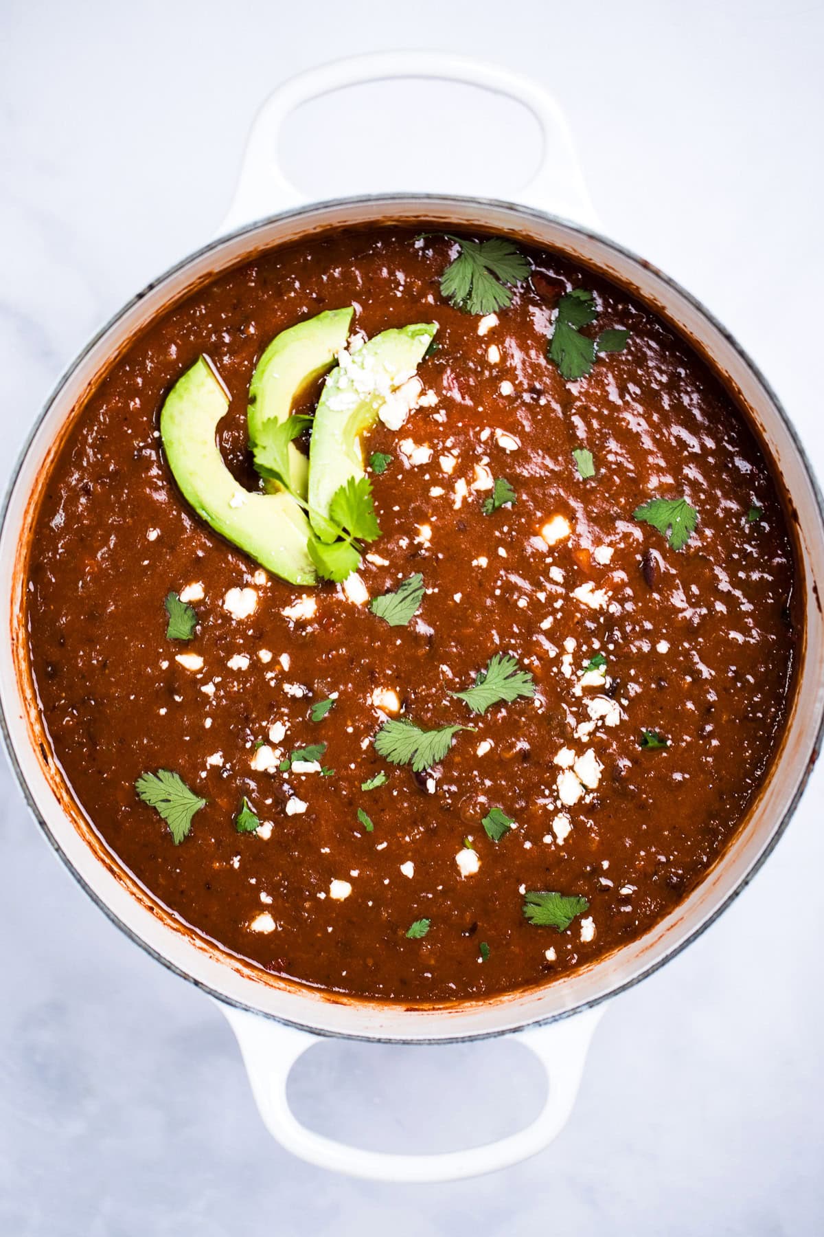 A dutch oven sitting on a table, full of black bean soup topped with queso fresco, fresh cilantro leaves, and avocado slices.