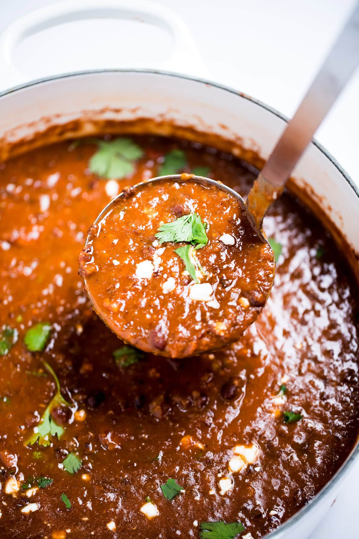 A ladle lifting black bean soup from a dutch oven, topped with queso fresco and fresh cilantro leaves.