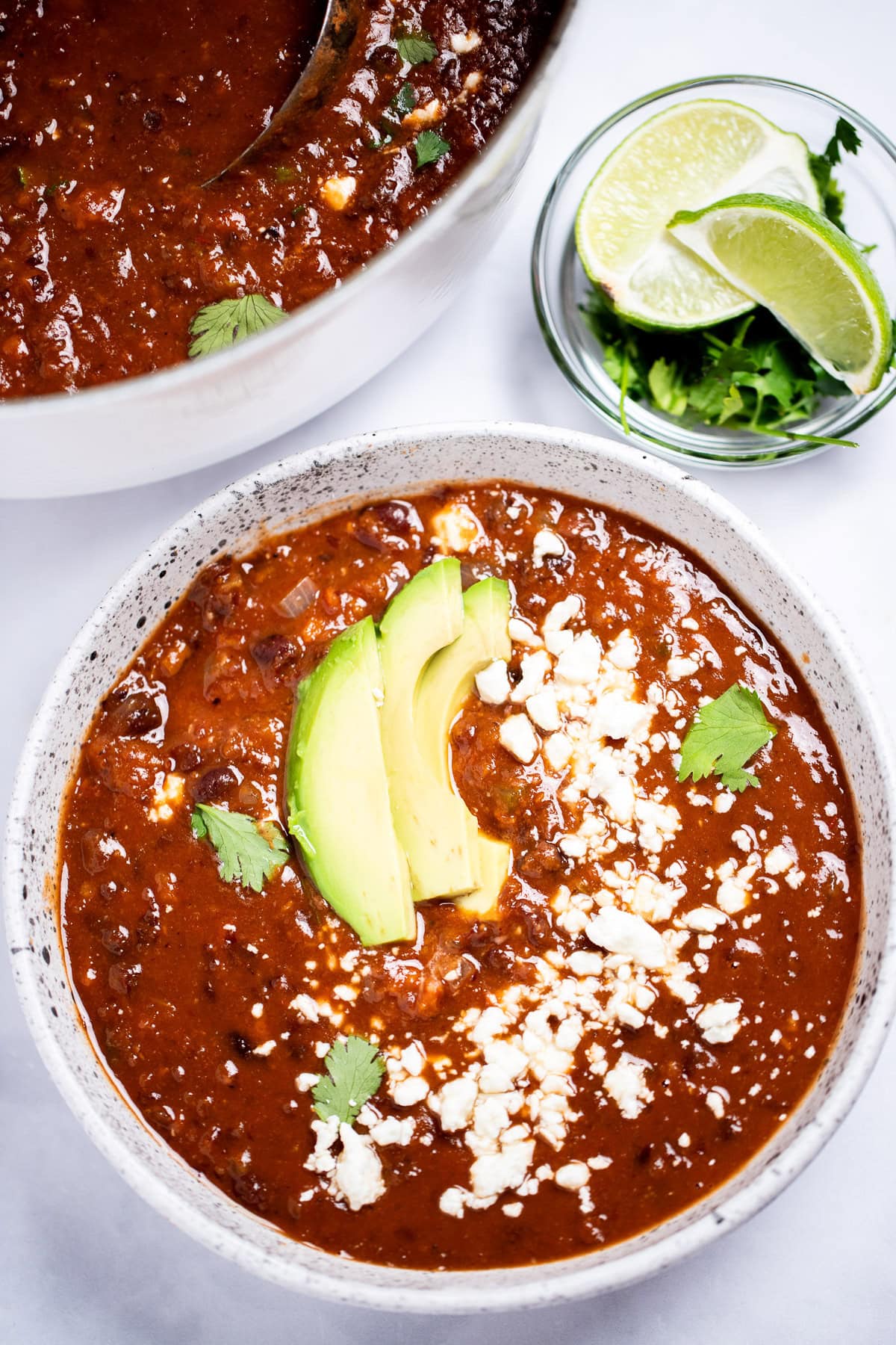 A bowl of black bean soup topped with queso fresco, cilantro leaves, and sliced avocado, with a bowl of cilantro and lime wedges, and a dutch oven of soup in the background.