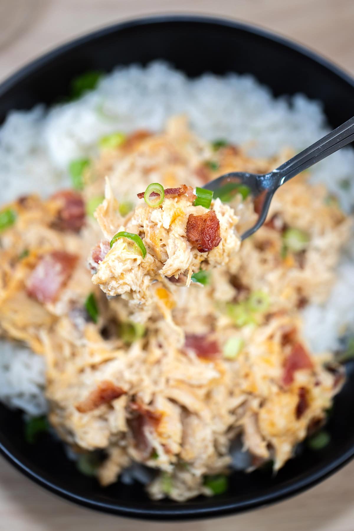 A fork lifting some cream cheese chicken up from a bowl of rice, topped with crispy bacon pieces and green onion.