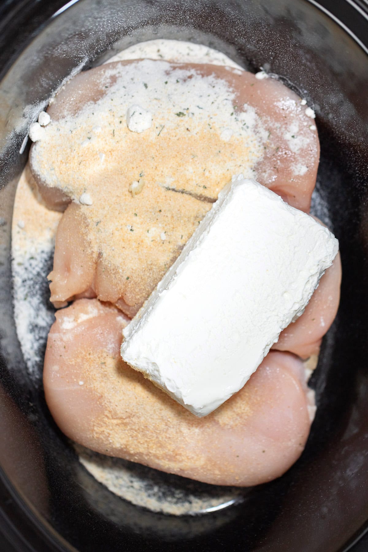 Raw chicken breast topped with seasoning and a block of cream cheese in a crockpot before being cooked.