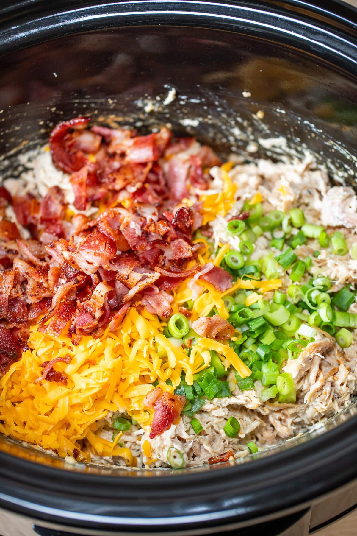 A crockpot with cooked cream cheese chicken shredded and topped with cheddar cheese, bacon, and green onions before being stirred together.