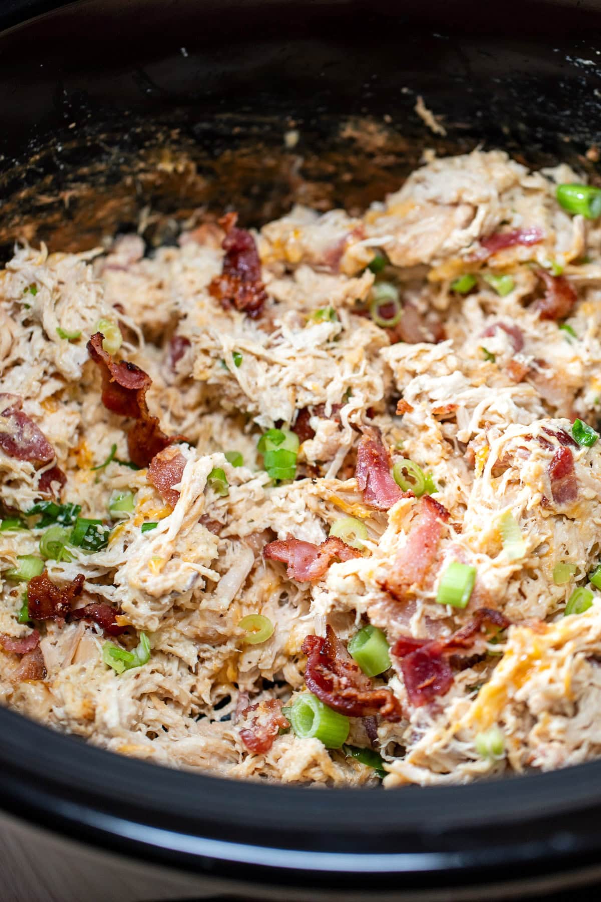 A crockpot with ready to eat cream cheese chicken mixed with crispy bacon, green onions, and shredded cheddar cheese.